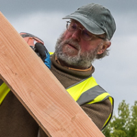 Man in a safety vest and cap lifting a wooden plank outdoors under a cloudy sky.