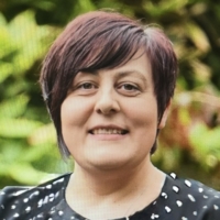 Smiling woman with short, dark hair, wearing a black top with white spots, standing in front of greenery.