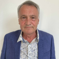 Older man with grey hair in a blue blazer and floral shirt stands against a plain white background.