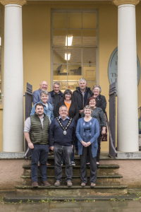 A group of eleven adults pose and smile together on stone steps outside a yellow building with columns.