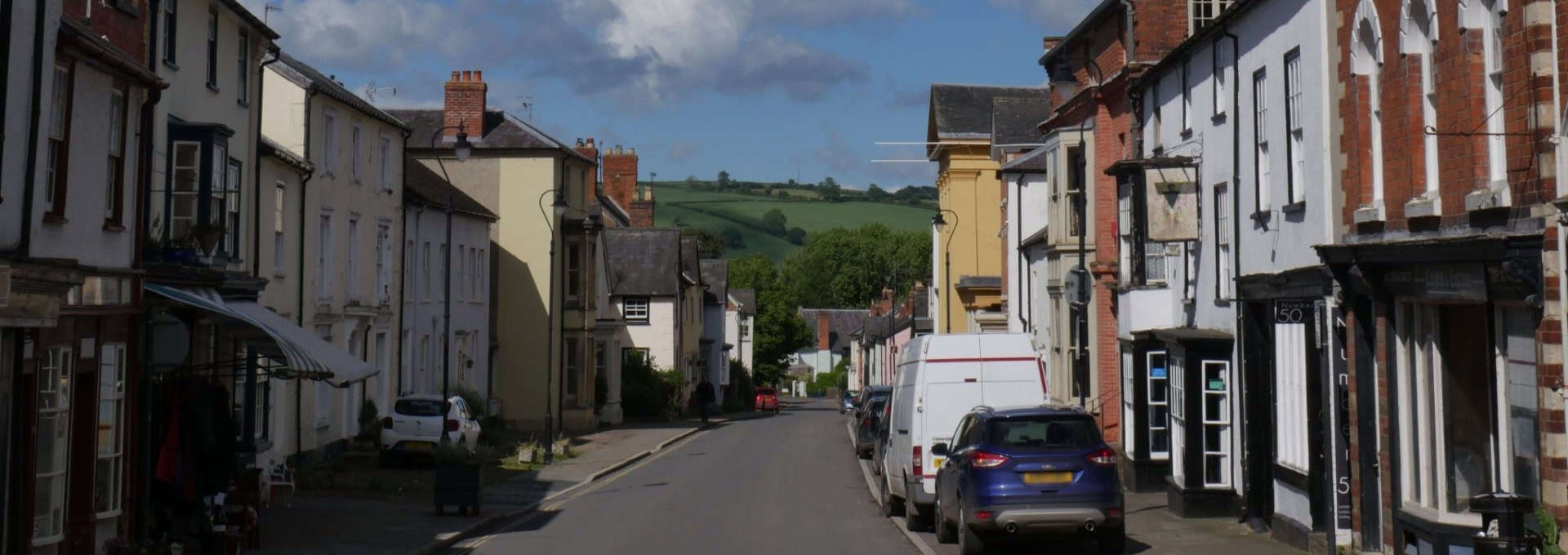 A quiet street in a small town with parked cars, shops, and green hills in the background under a blue sky.