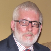 An older man with white hair and beard, wearing glasses, a suit, and a striped tie, sits indoors.