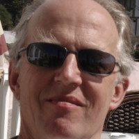 Man with greying hair wearing sunglasses, sitting outside on a sunny day.