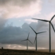 Three wind turbines stand on grassy land under a cloudy sky with sunlight breaking through.